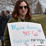 A woman holds a sign at a protest reading "Maine deserves care not cuts. Fund care. Not billionaires."