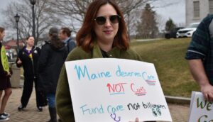A woman holds a sign at a protest reading "Maine deserves care not cuts. Fund care. Not billionaires."