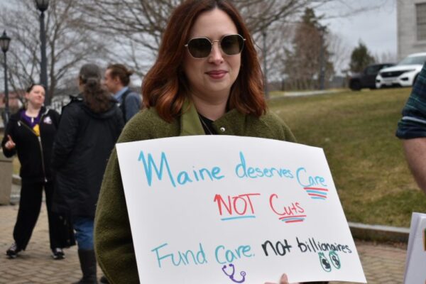 A woman holds a sign at a protest reading "Maine deserves care not cuts. Fund care. Not billionaires."