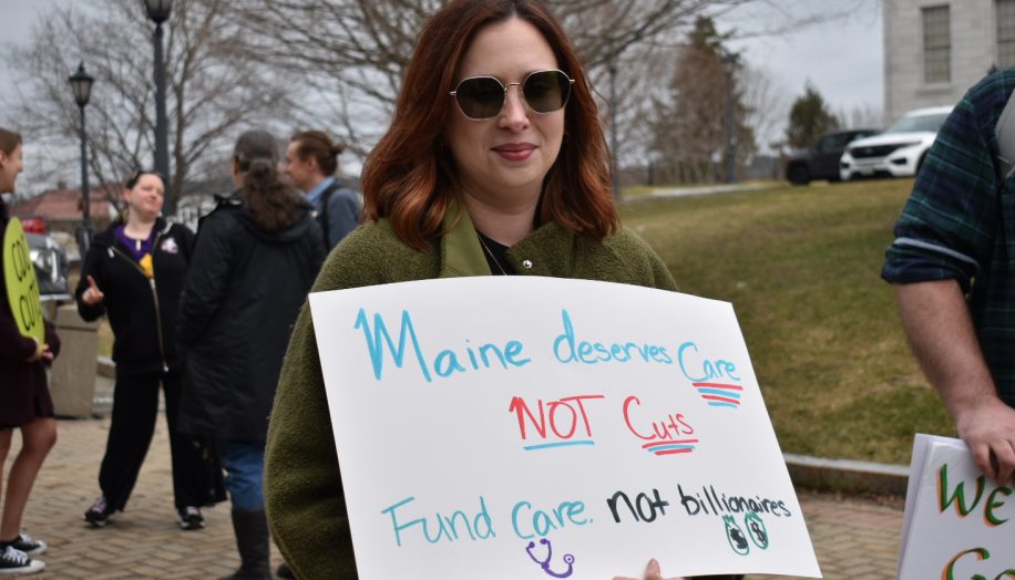 A woman at a protest holding a sign that reads "Maine deserves care not cuts. Fund care. Not billionaires."